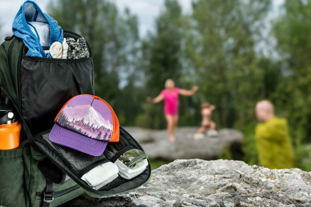 A backpack with hiking gear, including hats and water bottle, on a rock with hikers in the background.