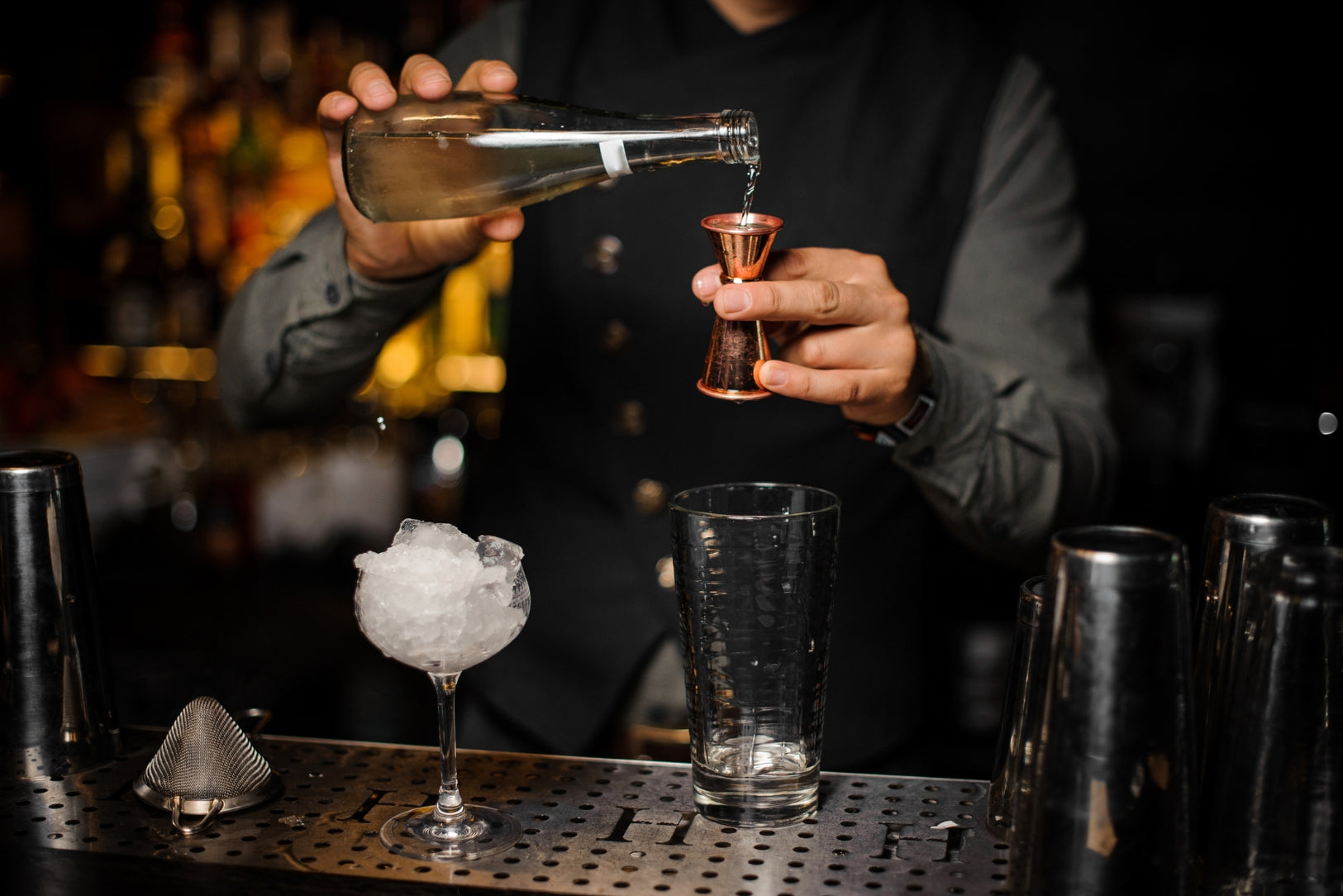 Bartender pouring a clear drink from a bottle into a copper jigger at a bar counter with glasses, ice, and cocktail tools in view